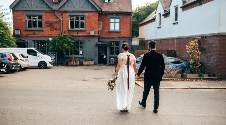 Man & woman seen from back in wedding attire walking toward pub